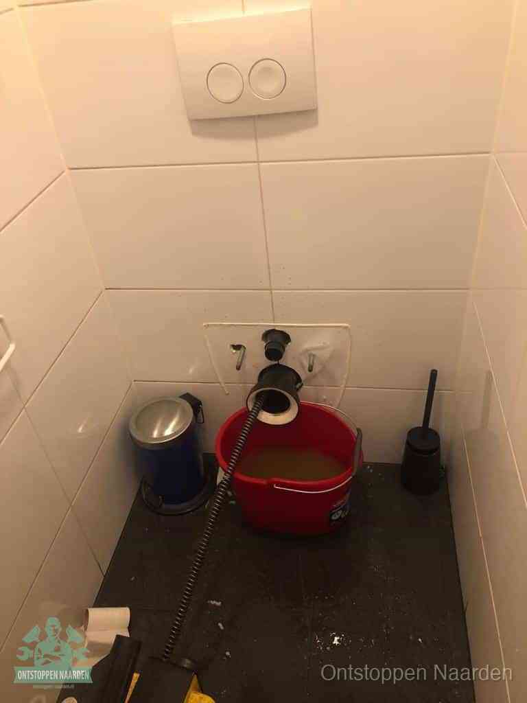 Een ontstopte wand-wc met verwijderde toiletpot waar een ontstoppingsveer in de wanduitgang verdwijnt, het achterhalen waar de verstopping zit in Naarden