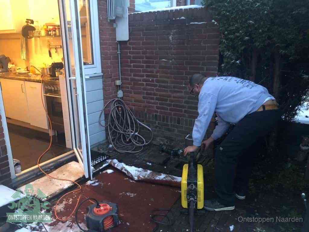 Een monteur werkt buiten bij een open riool-/grondput met een elektrische ontstoppingsmachine om een verborgen afvoerprobleem op te sporen in Naarden