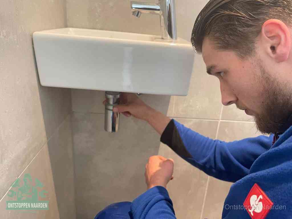 Een monteur draait met de hand de zwanenhalssifon onder een fonteinwasbak in de toiletruimte los om de bron van rioollucht in Naarden te vinden