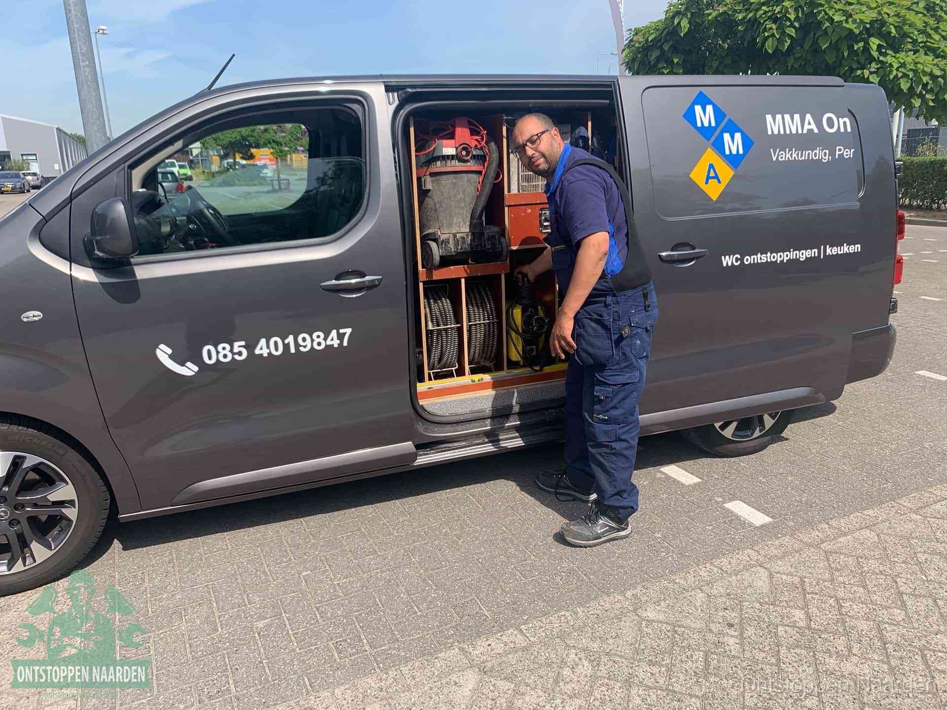Een monteur bij de open schuifdeur van zijn bedrijfsbus op een parkeerplaats, klaar om de oorzaak van terugkerende afvoerverstoppingen aan te pakken in Naarden