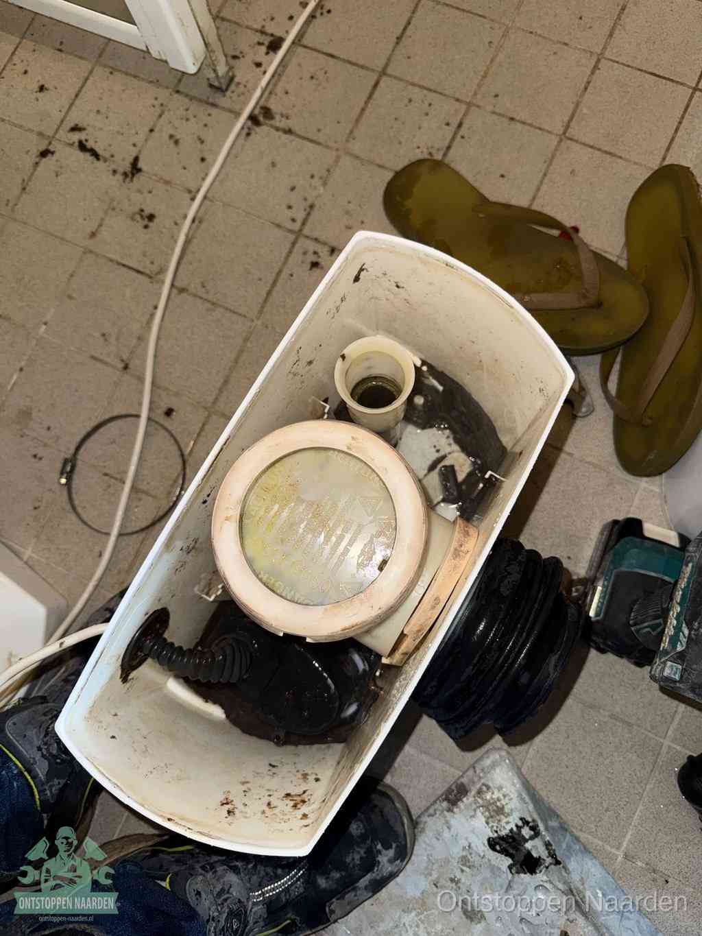 Een gedemonteerde toiletpot op de vloer met ontstoppingsveer en gereedschap ernaast, het verwijderen van een vastzittend voorwerp uit de wc in Naarden