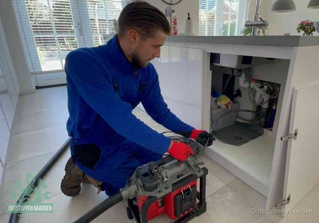 Een monteur voert een professionele elektrische ontstoppingsmachine in de afvoer onder de spoelbak tijdens het gootsteen ontstoppen in Naarden